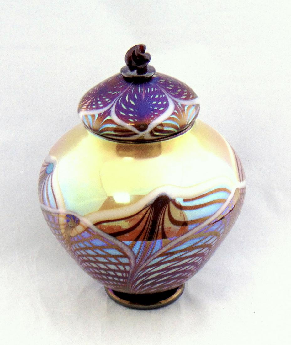 Rick Strini Saturn glass Jar: Ovoid jar in caramel glass decorated with a ruby and white pulled feather, fitted with a matching lid with twist finial, signed Strini 02, 11" ht. Condition: good with no defects found.