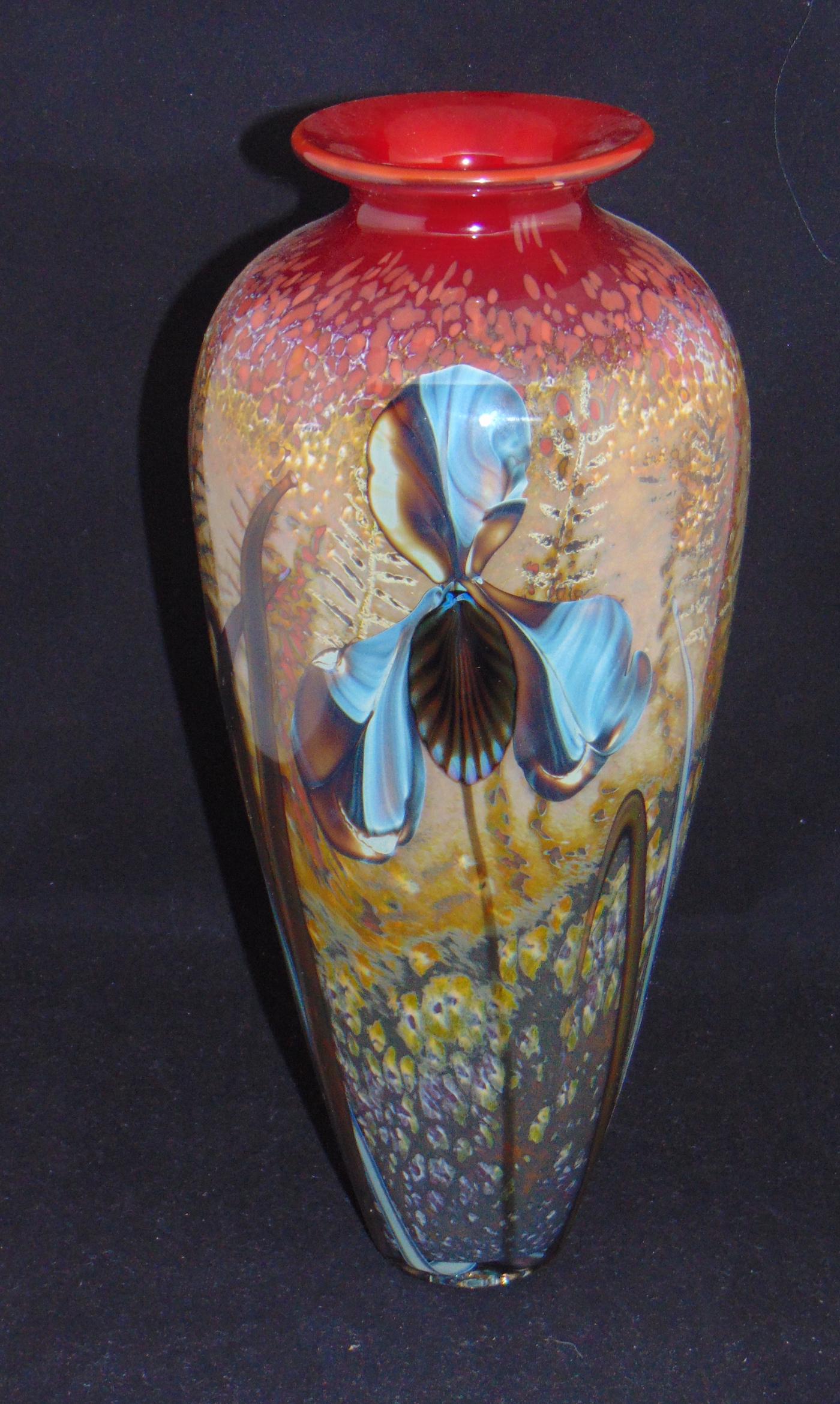 Rick Satava "Iris in Field" glass vase: Cylindrical form in mottled red and brown glass decorated with a long stem blue Iris, signed Satava 00, 14 1/2" ht. Condition: good with no defects found.