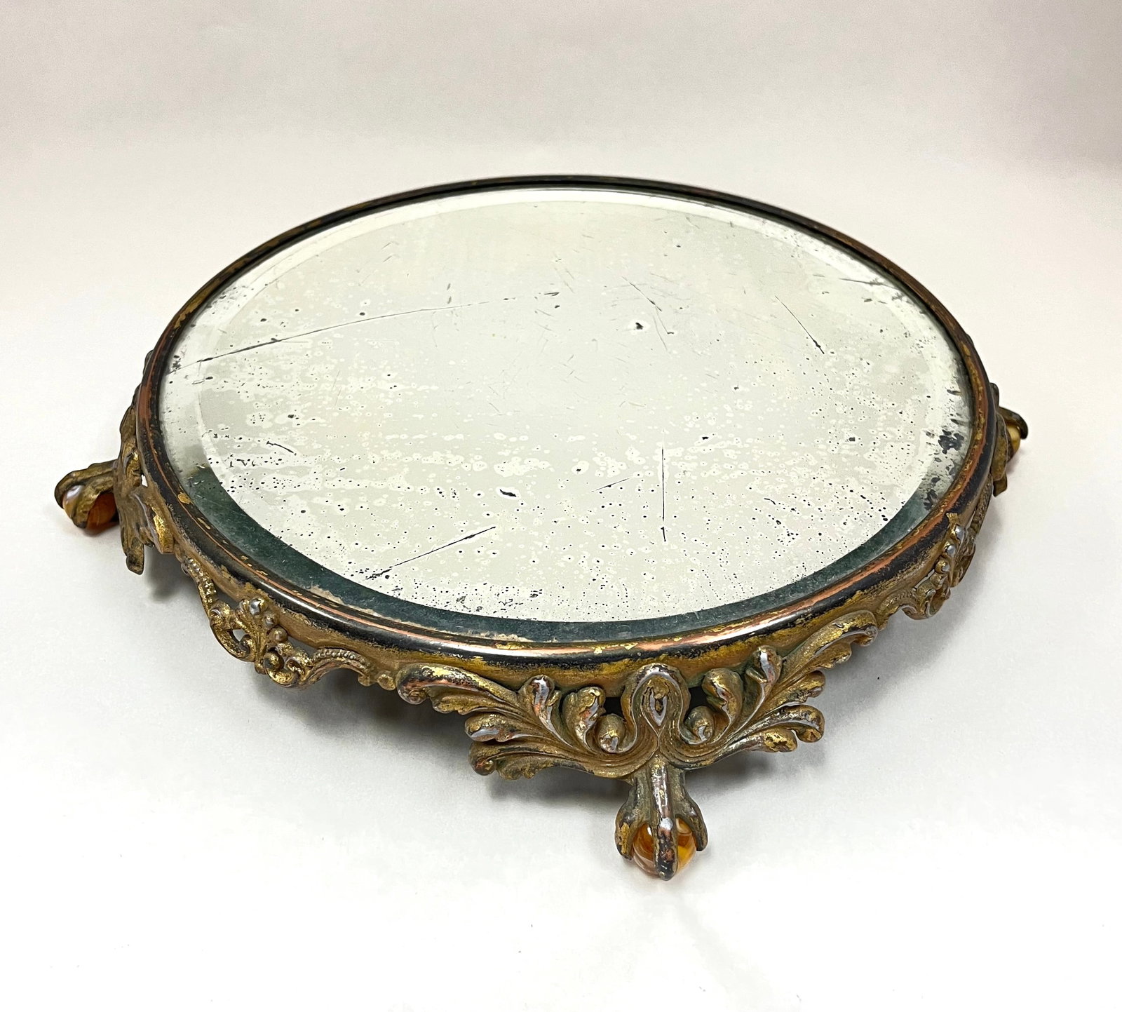 Glass and metal Plateau: Beveled edge ovoid mirror set in a claw foot metal stand with embossed acanthus leaves, each foot holds a marble, 11" wide. CONDITION; wear to the mirror, no chips, cracks, repairs or material defects
