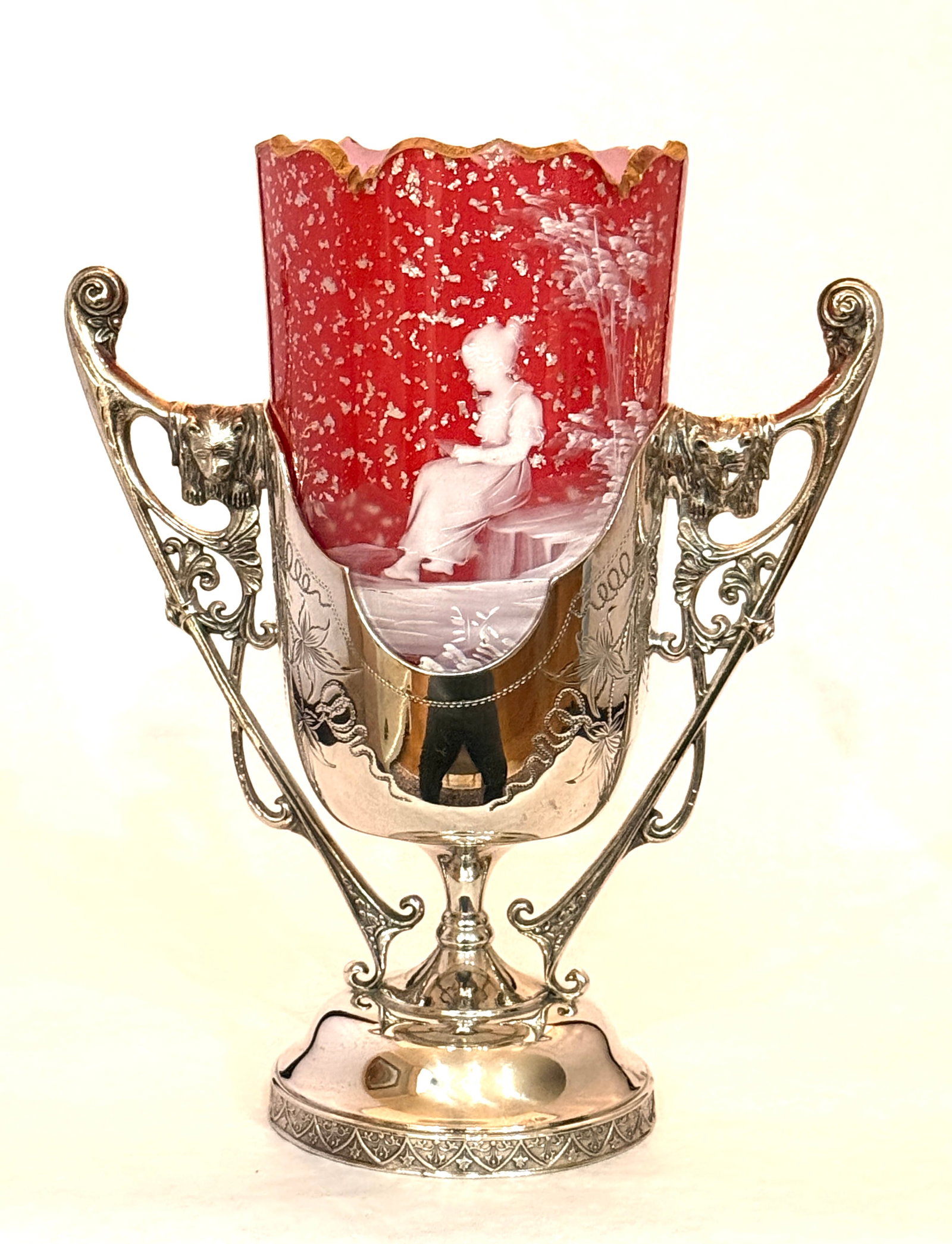 Mary Gregory Celery Vase: Mary Gregory cranberry cased insert with a gold rim decorated with a Victorian lady on a bench and overall internal silver flecks, set in an Aurora caddy with lion mask handles, 9 1/2" ht. CONDITION:
