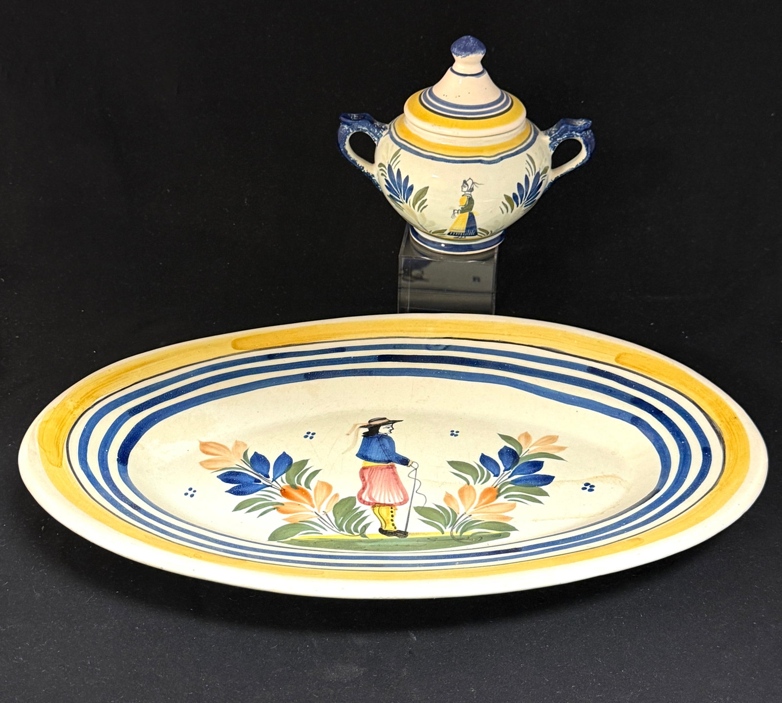 2 HenRiot Quimper Items: 2 HenRiot Quimper yellow and blue banded dinnerware items with Breton figures including a platter and a covered sugar bowl; length 15". CONDITION: Quimper has propensity to have minor chips, glaze