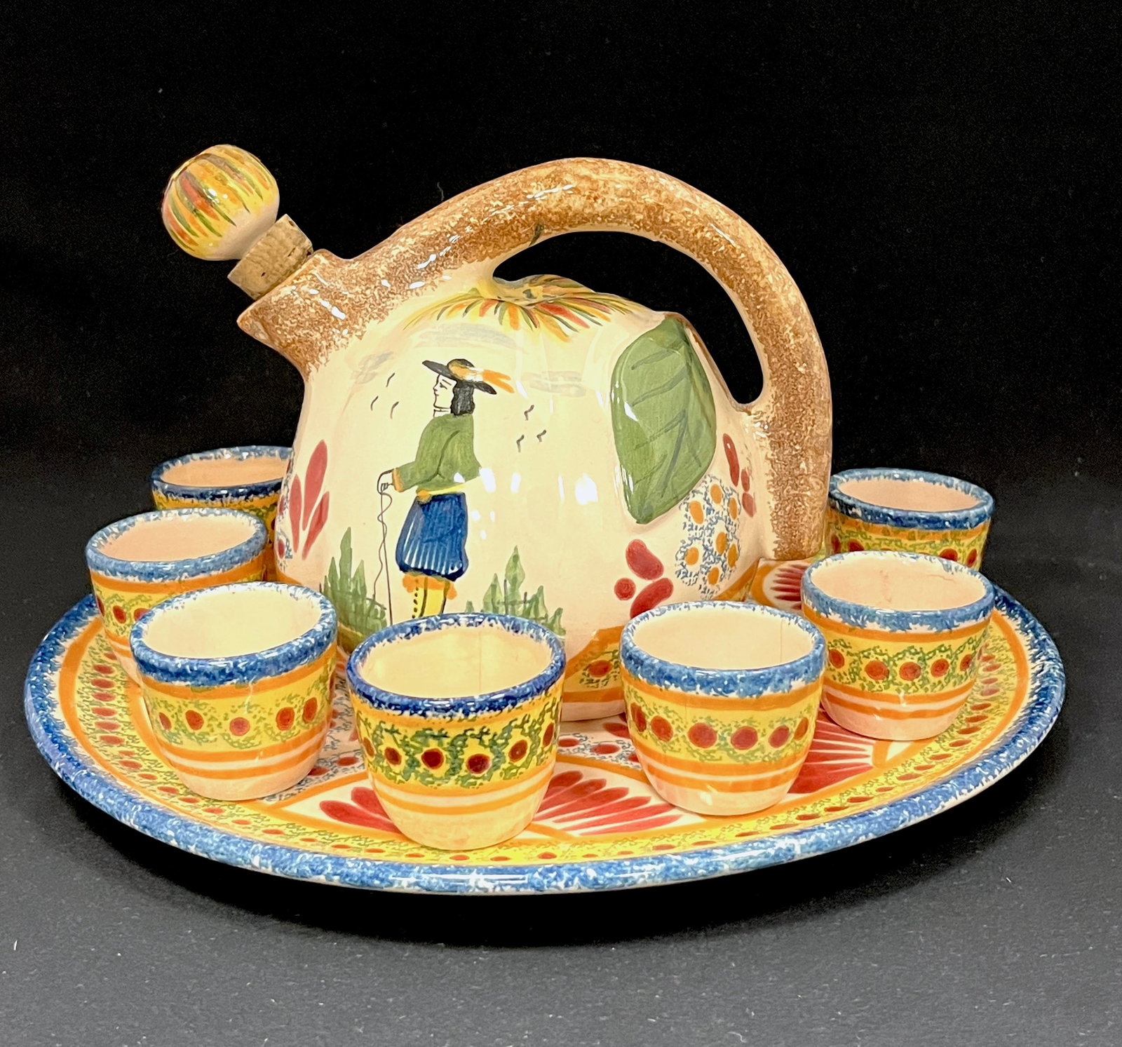 HenRiot Quimper Cider Set: Henriot Quimper Cider set decorated with Breton with walking stick and a sponge a la touch? decor, 10" wide. CONDITION: two cups with hairlines. Quimper has propensity to have minor chips, glaze