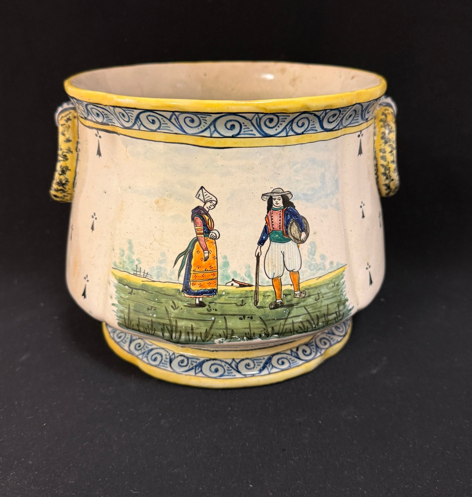 HB Cache Pot: HB cache pot flanked with dimensional rings, back and front decorated with Breton couples in a country setting, Fleur de Lis, ermine tails and a two toned scrolling borders decorated rim and base;