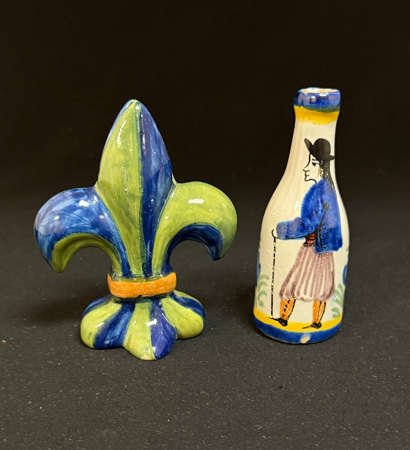 2 Quimper Items: 2 Quimper items an HB Fleur de Lis menu holder and a petite bottle with Breton using a walking stick; ht. 3 1/2" circa 1900. CONDITION: Quimper has propensity to have minor chips, glaze loss/wear and