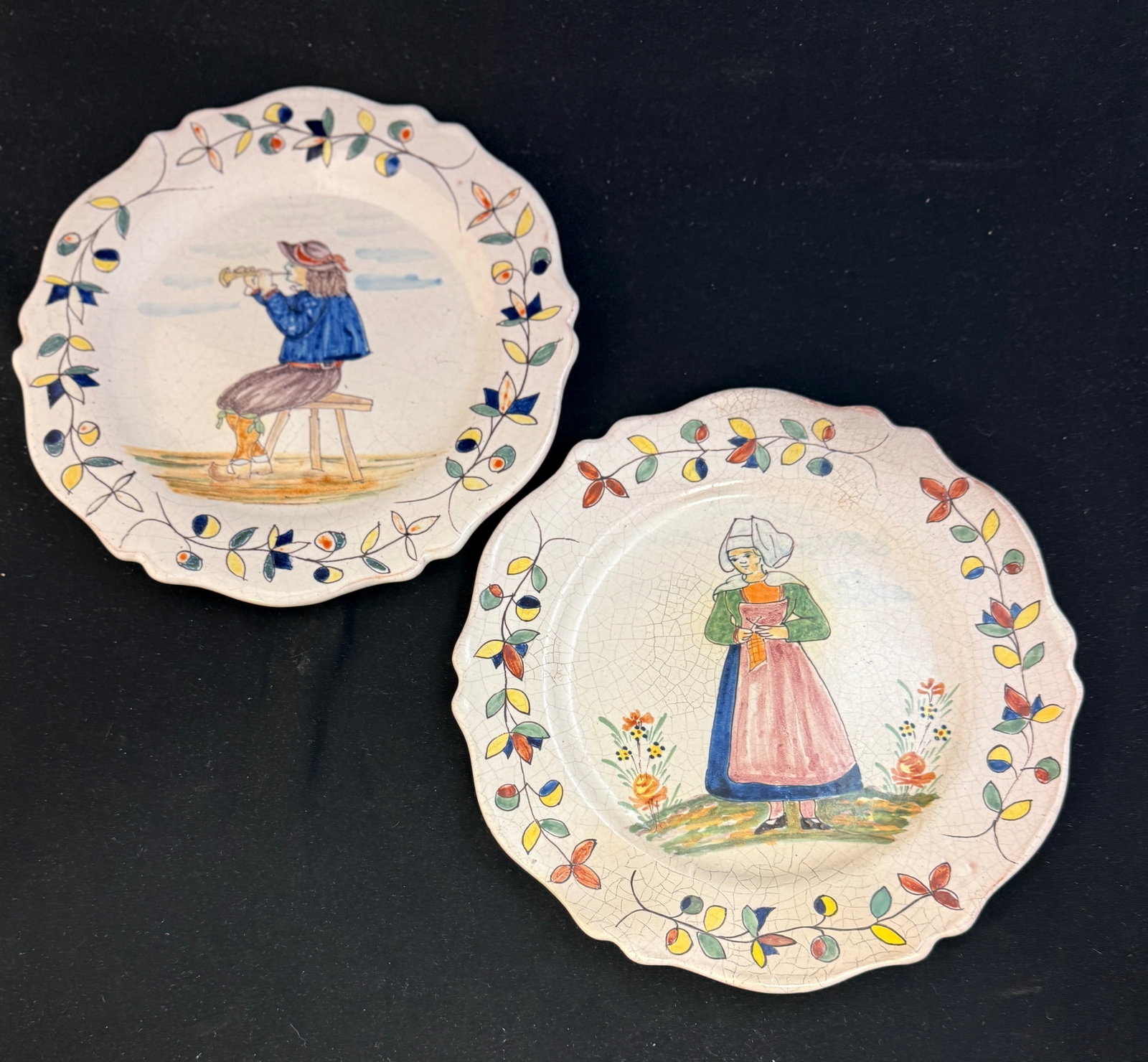 2 Malicorne Plates: 2 Malicorne plates with traditional border, one with seated Breton playing a bombarde the other with a young Bretonne wearing an apron and coif holding knitting; diameter 9 1/2" circa 1895-1920.