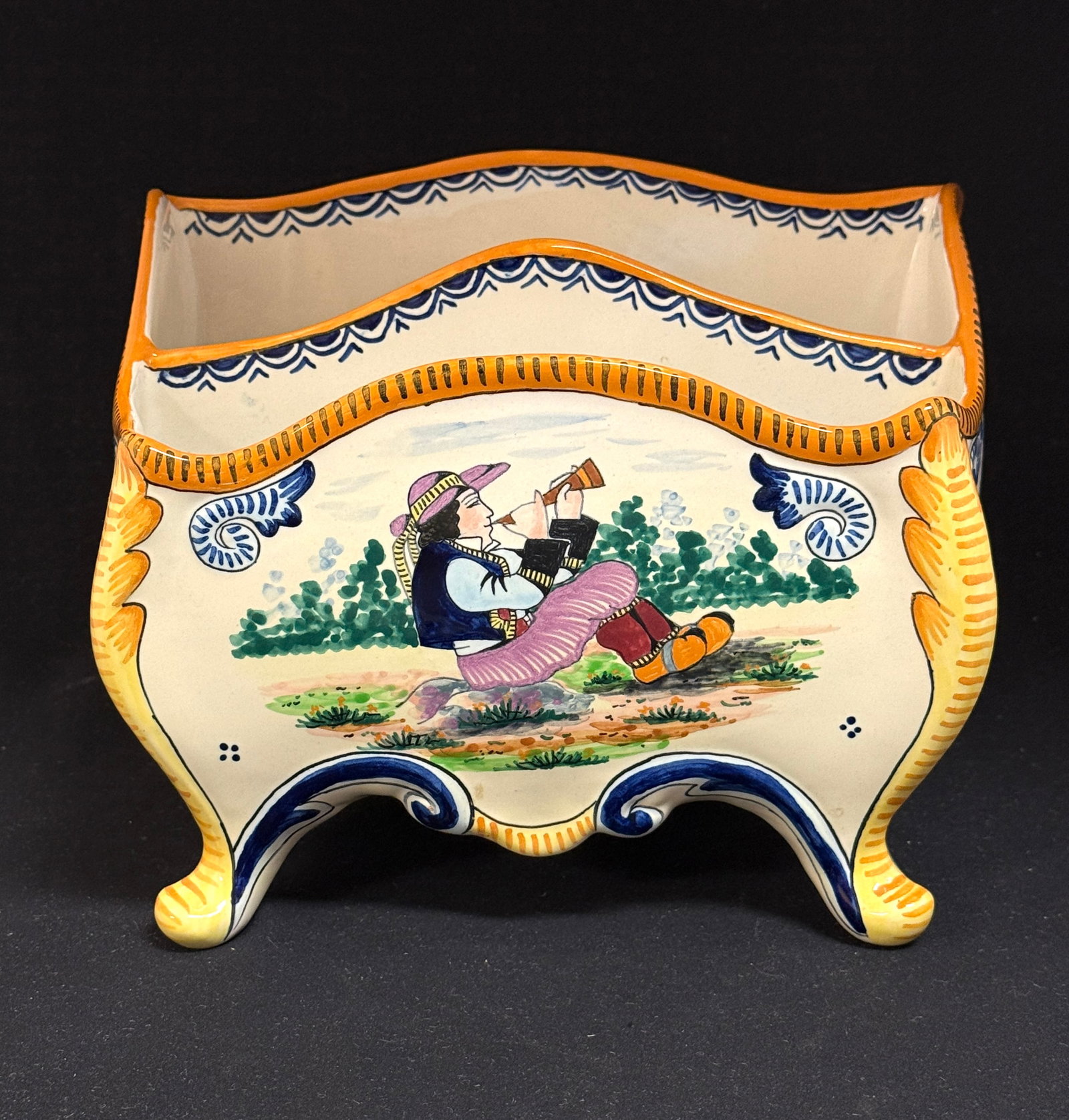 HB Quimper Footed Jardiniere: HB Quimper footed chest form jardiniere/letter holder with young Breton playing a bombarde resting on a rock with tone on tone flourishes, 4 dot motif, ermine tails, a large Fleur de Lis, and ajonc; h