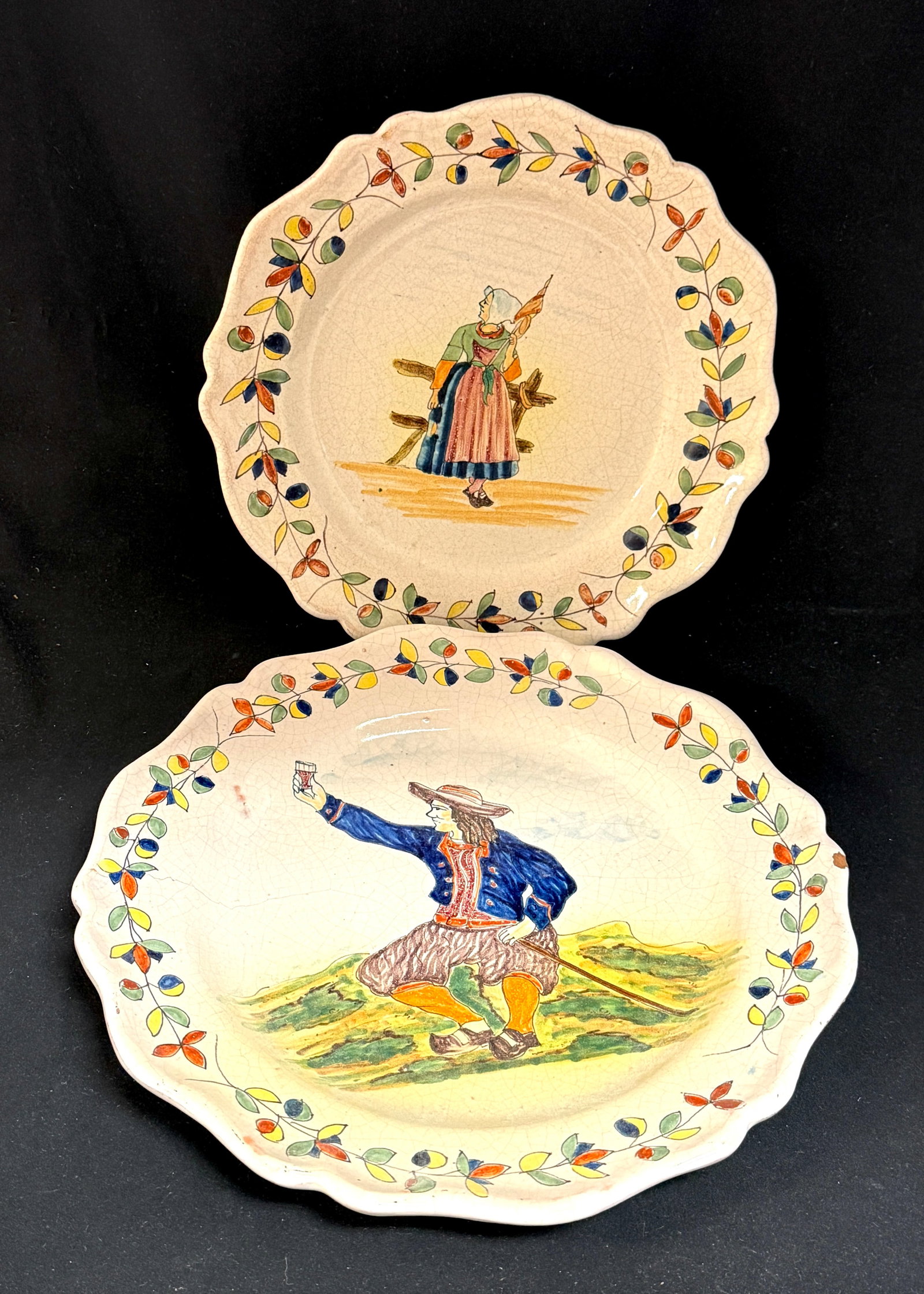 2 Malicorne Plates: 2 Malicorne plates with traditional borders one with Bretonne holding a staff leaning on a fence, the other a seated Breton holding a walking stick and raising his glass in a toast, marked PBX;