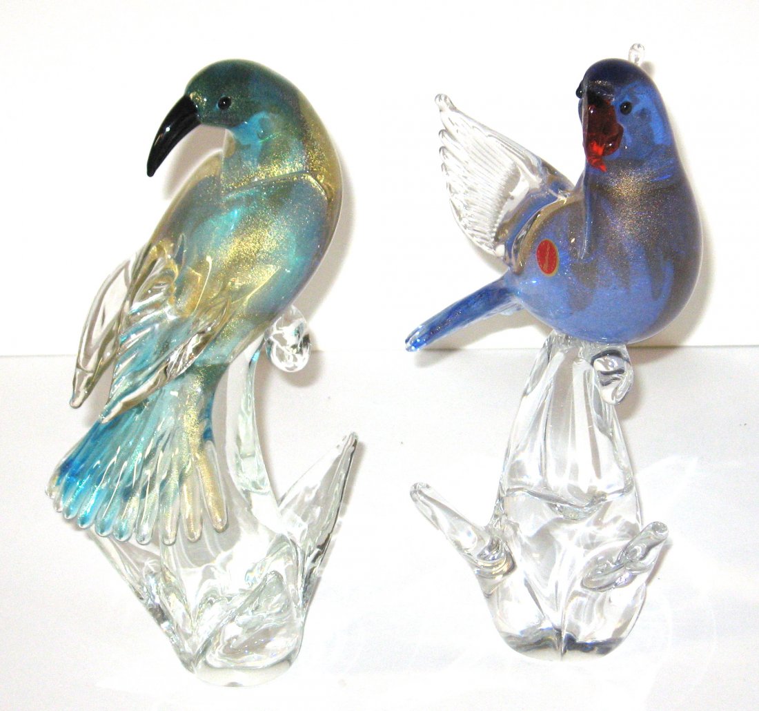 Two colorful Murano exotic birds,: The first a dark blue winged parrot with red beak, the other a teal bird with black beak, both sitting on glass rocks and adorned with gold mica, 11" ht.