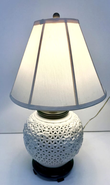 Blanc De Chine Reticulated Cherry Blossom Lamp: A white ceramic table lamp featuring a reticulated, globular Blanc De Chine porcelain base with raised cherry blossom designs, set on a dark stand and topped with an off-white fabric shade, it is 17 i