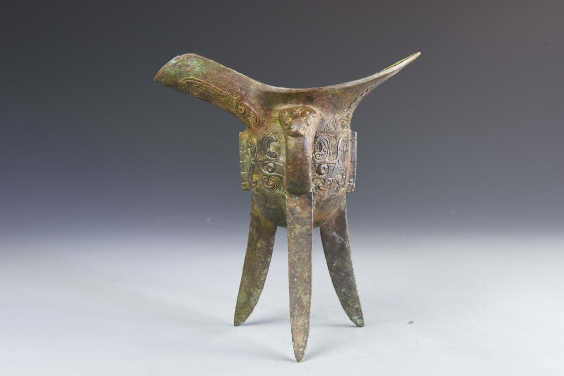 Chinese Bronze Jue: China, antique bronze Jue, body with two high swooping sides, three tall legs and rounded handle, lightly carved details around exterior. Height 8 in.