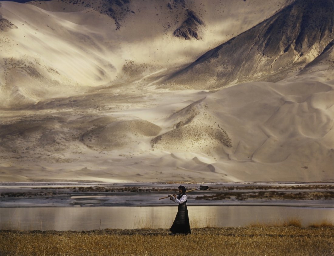 Photograph, Lisa Kristine: Photograph of a woman with a shovel slung over her shoulder, she walks by a small pond with large snowy mountains in the background, by Lisa Kristine, signed, titled, and dated.