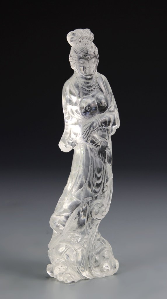 Chinese Crystal Guanyin: China, carved crystal figure of Guanyin, in a standing position and crossed arms, wearing flowing detailed robes with exposed breasts, standing on an organic formed base. Height 7 7/8 in.