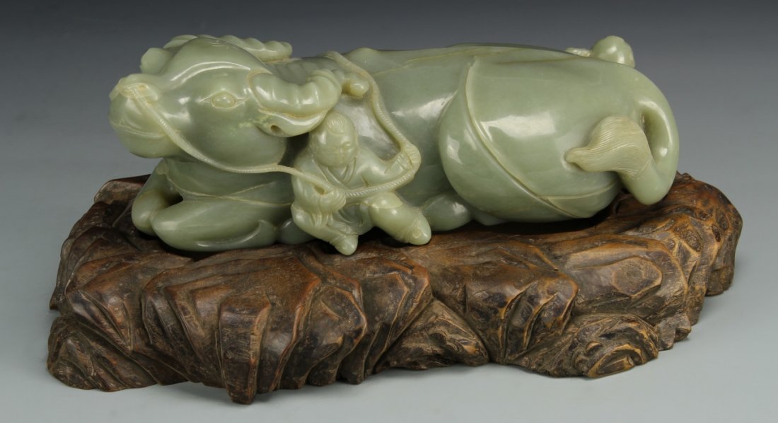 Chinese Large Celadon-Green Jade Cow and Boy: China, 19th C., a boulder carved in the form of a recumbent cow with two boys, one sitting next to the cow, holding a rope and the other standing at the back, holding a Ruyi scepter, with carved hardw