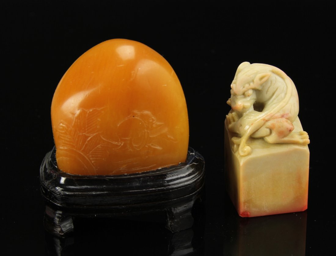 Two Chinese Shoushan Seal Chops: China, two Shoushan seal chops, one in a deep orange hue on a wood stand, and the other in rectangular form, with carved mythical animal on top. Height 2 in.