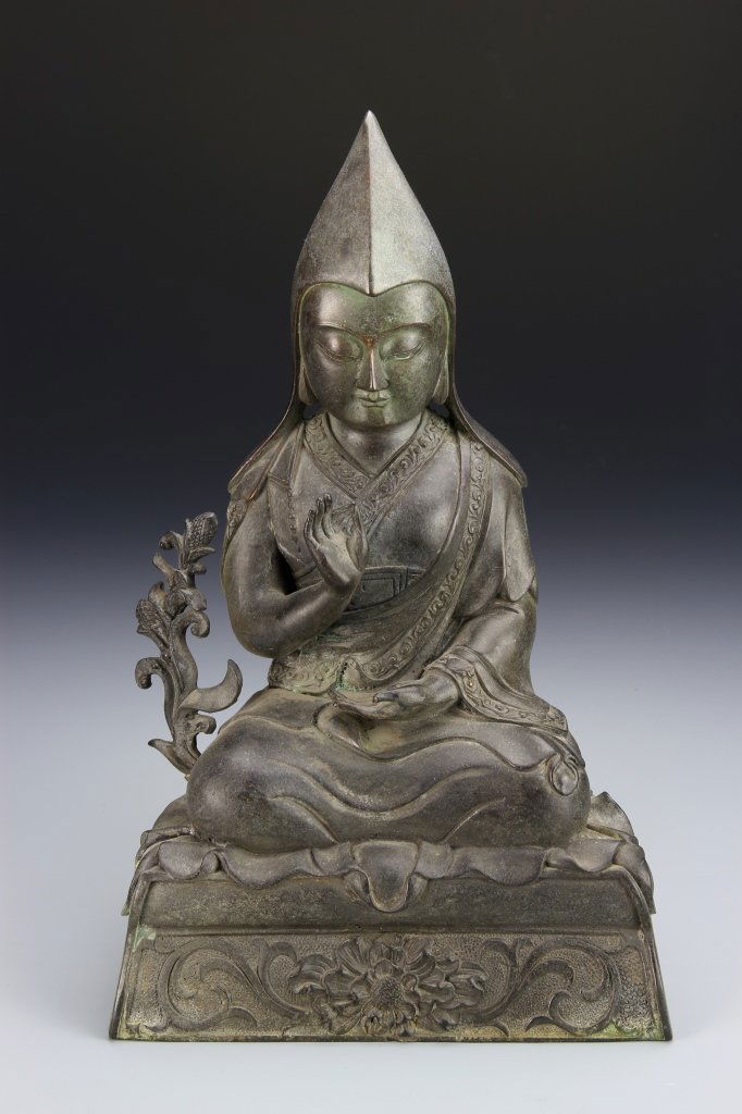 Tibetan Bronze Seated Buddha: Tibet, bronze figure of Buddha, in a seated position, with patterned robes, a serene facial expression, with a plant at the side, on a raised pedestal base. Height 10 1/2 in.