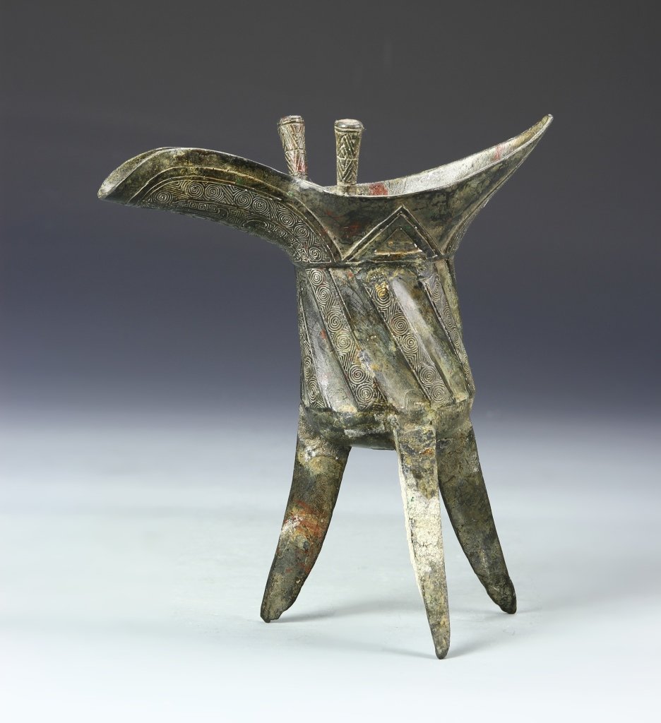 Chinese Bronze Wine Cup: China, bronze wine cup, cylindrical form with a flared rim and an extended open spout, standing on three legs, intricately incised with swirling patterns and geometric designs, with patina. Height 7 3