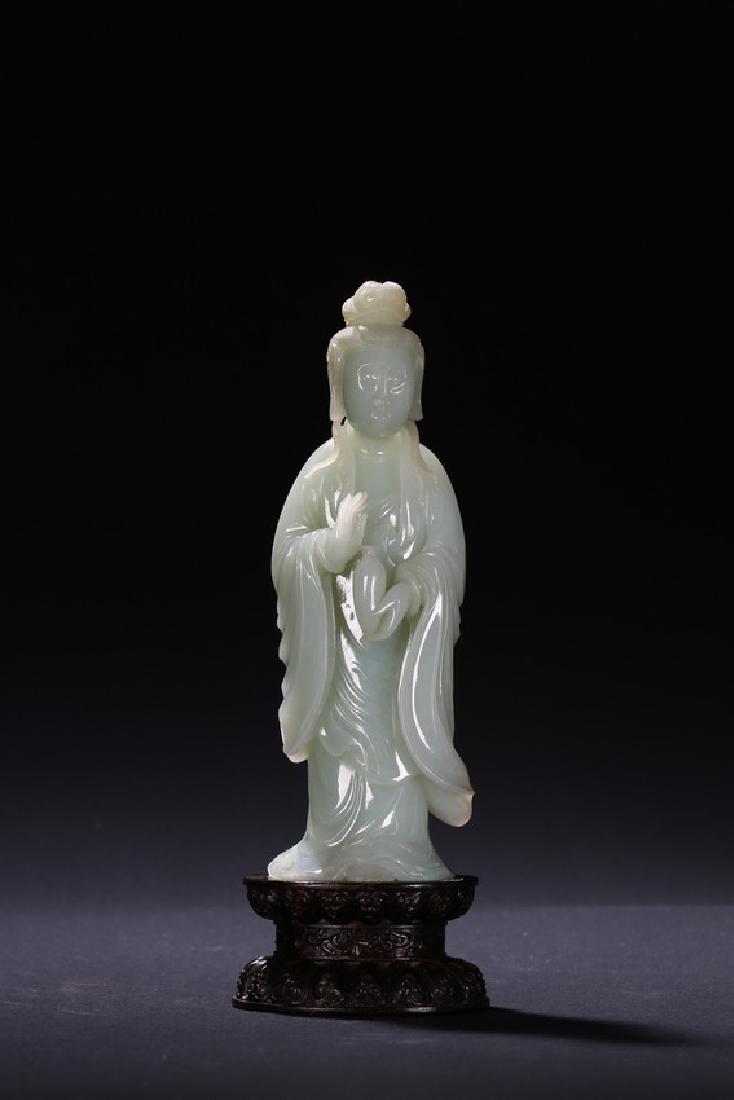 A WHITE JADE FIGURE OF STANDING GUANYIN: The Guanyin is shown standing with left hand holding vase and right hand raised in mudra, wearing voluminous robe falling in elegant folds, face with serene expression, all supported on a wood lotus s