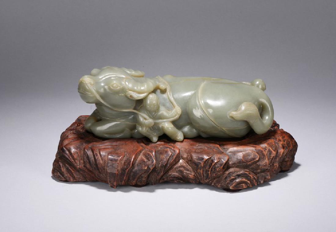 A CELADON JADE CARVING OF BUFFALO AND BOYS: The recumbent buffalo is carved with four feet tucked underneath body,the head slightly raised and tail swept to the left,with a boy sitting beside holding a rope tied through its nostril and over its