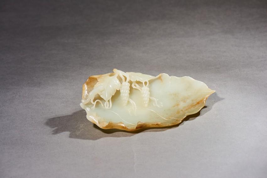 A WHITE AND RUSSET JADE LEAF BRUSH WASHER: Of leaf-shape, carved to the interior in relief with clusters of grapes and squirrel, the stone is of polished white tone with russet patches and inclusions.Qing Dynasty period. Length: 7 1/8 in (18.0