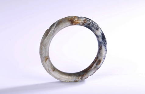 A JADE CARVED 'CHILONG' BANGLE: Carved in relief with a sinuous chilong around the bangle, the stone is of mottled white, black, and russet tone. Ming or Qing Dynasty period. Inner diameter: 64 mm Thickness: 13mm Provenance: From th