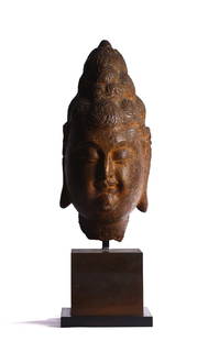 A LARGE CHINESE ARCHAIC STONE BUDDHA HEAD: The Buddha's face is carved in a full-frontal view, featuring prominently hooded eyes, a small mouth, and a distinct brow. This face is framed by elongated ears with dangling earlobes, while the hair