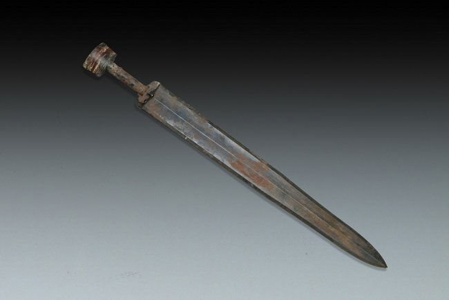 A CHINESE ARCHAIC BRONZE SWORD: This bronze sword has a short hilt with a rectangular pommel at the end. The sword guard slightly extrudes outward from the flat side of the blade, with an angular tip. The blade edge is thin, allowin