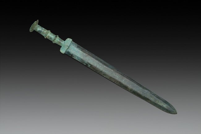 A CHINESE ARCHAIC BRONZE SWORD: This bronze sword had a thin handle with two extruding rings and a small flat pommel on the bottom. Sword guard had intricate carvings of patterns. Han Dynasty period. L: 14 5/8 in (37 cm)
