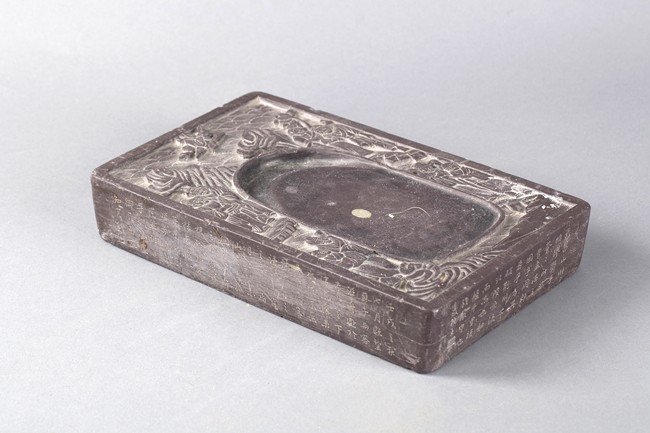 A CHINESE CALLIGRAPHY AND ELDER INKSTONE: This well-carved Chinese inkstone has many Chinese characters engraved on all four sides. The top is decorated with images of elderly figures on the edges. The back is carved with an elder and his att