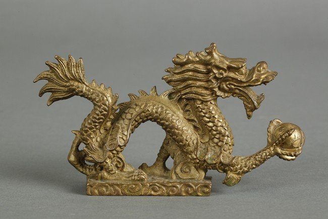 A BRONZE DRAGON DECORATIVE FIGURE: This bronze dragon is in golden color with its ferocious mouth open. The dragon clutches an orb with its five claws, while traversing through the clouds. Dimension:H: 2 3/4 in (7 cm) W: 4 1/2 in (11.5