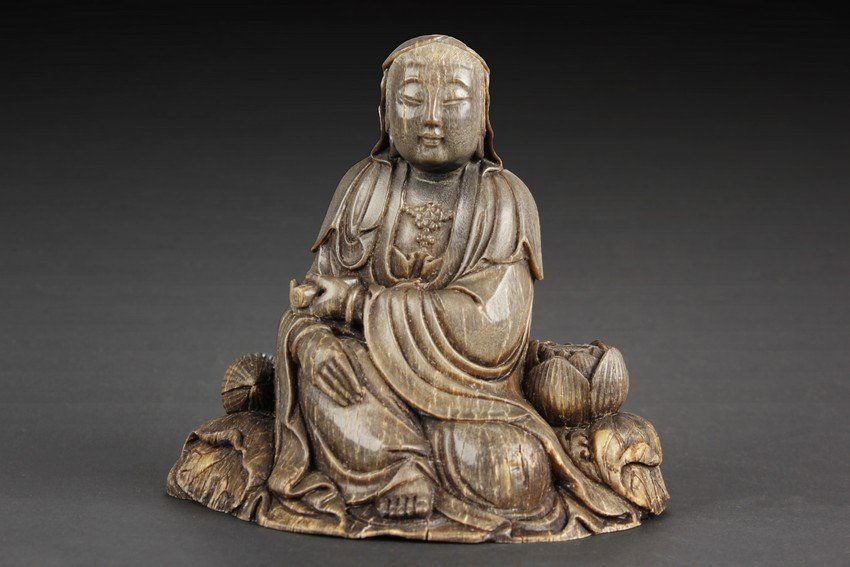 A RHINOCEROS HORN CARVING OF GUANYIN: Seated in Rajalatitasana with a serene expression, dressed in loosely draped robes left open at the chest to expose a decorated necklace, the face cast with serene expression, holding a rolled scroll