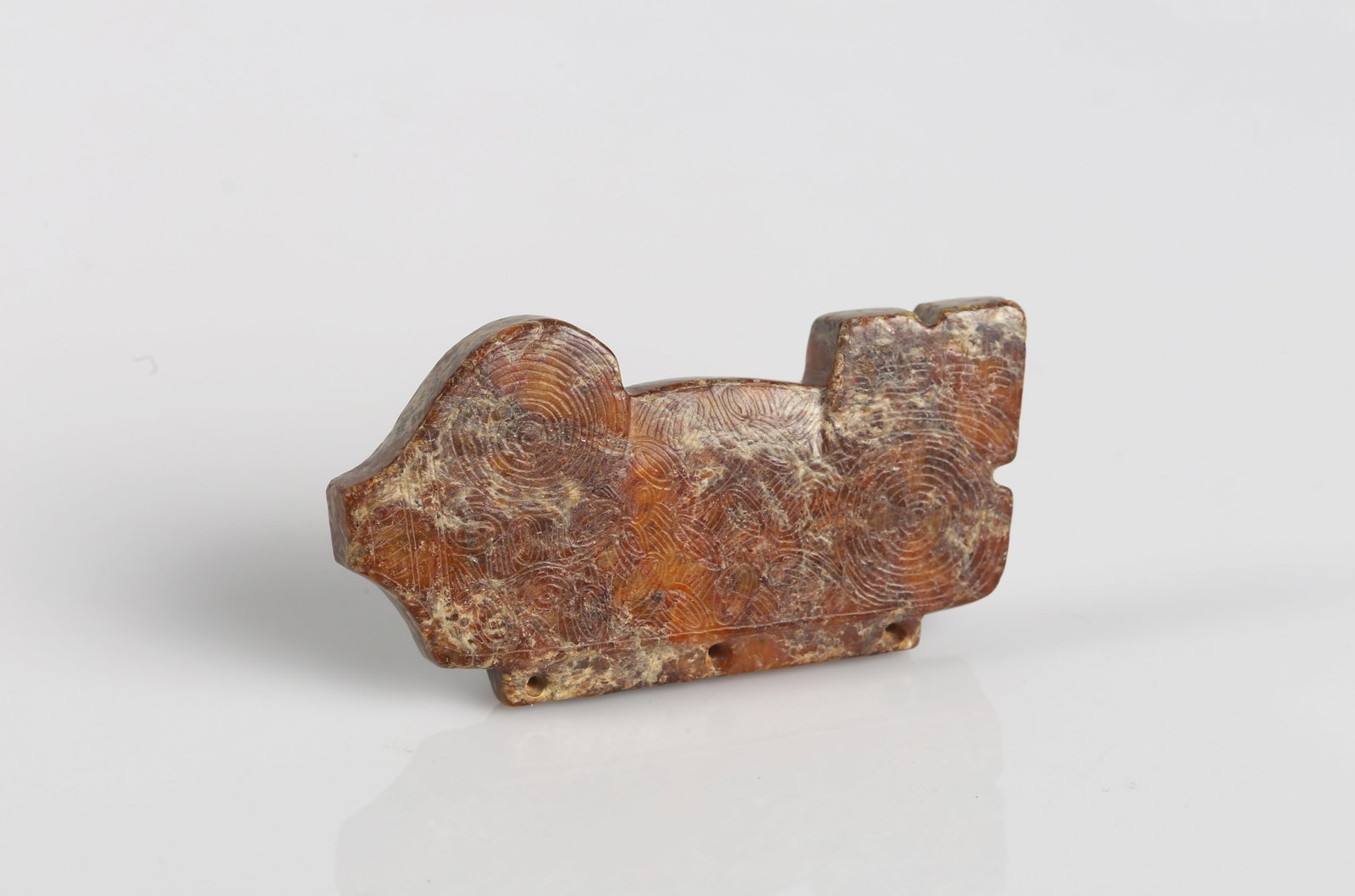 A Red Jade Carved 'Beast' Pendant: 文化期 玉豬形飾 Carved as a stylized recumbent creature, the flattened body incised with concentric scroll motifs, the stone of mottled russet tone; pierced