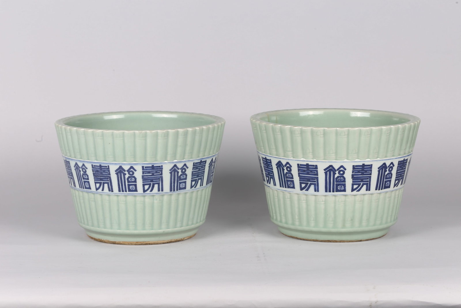A Pair of Celadon Glazed Blue and White Jardinieres: 清 豆青釉青花壽字紋花盆一對 Each jardiniere of sturdy cylindrical form with a wide flaring rim, the exterior applied with