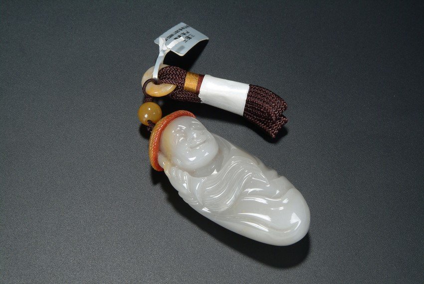 A CARVED JADE RIVER PEBBLE PENDANT: A pendant carved out of a jade river pebble in the form of a human figure. The carver used the natural color of the pebble skin to depict a straw hat on top of the figure. Qing Dynasty period. Dimensi