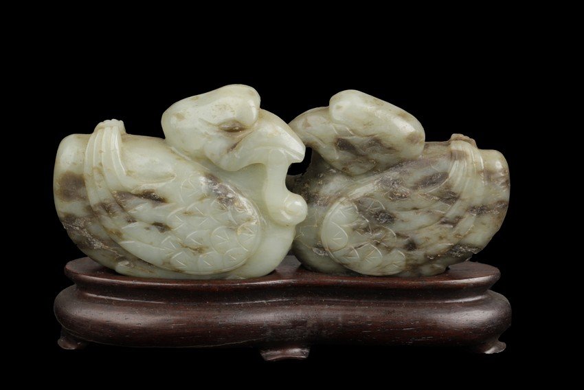 A celadon jade carving of two mandarin ducks: The figure depicts two mandarin ducks, wings on the side, swimming facing each other in close proximity. The stone of pale celadon color, with dark and brown markings. Includes stand. Ming Dynasty per
