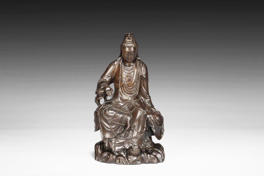 A ZITAN WOOD CARVED SEATED GUANYIN FIGURE: Seated in lalitasana on top of a wooden trunk, draped in long robe flowing freely with chest slightly exposed showing ornate necklace, right hand rests on an extended wooden armrest, left hand rests o