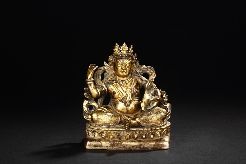 A TIBETAN GILT-BRONZE FIGURE OF YELLOW JAMBHALA: This gilt-bronze Tibetan deity is seated in lalitasana on a raised lotus throne, dressed in ornate clothes, completed with decorated headdress, earrings, necklace, and arm bands. He holds a golden rat