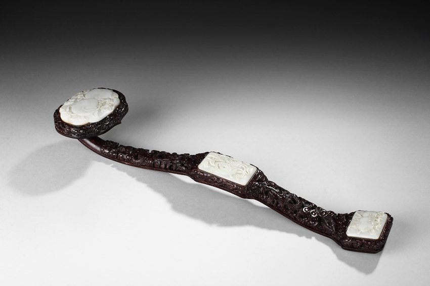 A CARVED HONGMU WOOD RUYI SCEPTER INSET WITH WHITE JADE: The ruyi scepter was carved from Hongmu hard wood, relief carved with dragons flying amidst clouds, with natural dark red color and grains. The head inset with a carved jade depicting peaches and bats