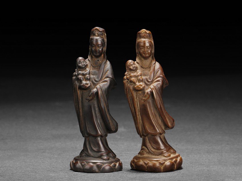 TWO WOOD CARVED GUANYIN FIGURES: The two Guanyin figures are almost identical in design, both stands on a lotus pad, with one hand holding a baby, symbolizing fertility. One Guanyin with brown color, the other with deeper brown-black