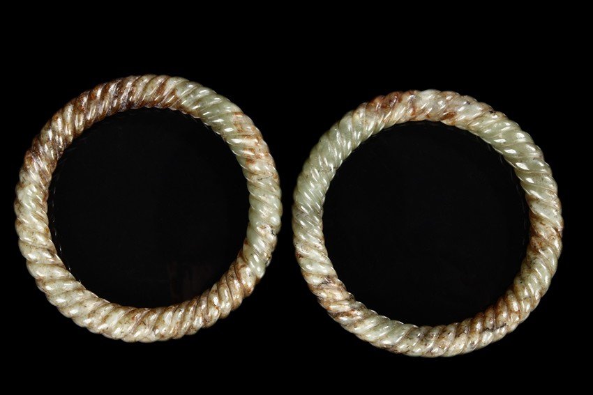A PAIR OF SPIRAL JADE BANGLES: Of round shape, both bangles carved in spiral on the exterior, the jade of celadon green with russet. Ming Dynasty period. D: 3 1/2 in (9 cm) 64g each