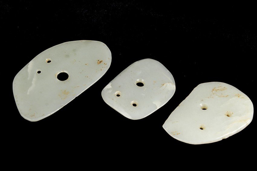 A GROUP OF THREE SMALL WHITE JADE PLAQUES: Each white jade shaped irregularly, with two smaller hole and one large hole in the center in triangular shape; the stone of translucent white color, with a little russet. Ming Dynasty period. Largest