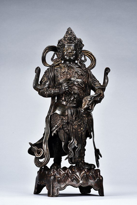 A PARCEL GILT BRONZE FIGURE OF VAISRAVANA: Cast standing upon a rockwork base, the figure is shown wearing elaborate chainmail completely covering the body and extending down to boots, left hand holding a mongoose, the right raised as if to ho