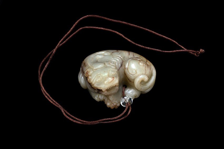 A WHITE AND RUSSET JADE 'BUDDHIST LION' CARVING: The mythical beast is shown seated on its haunches with heavy paws, its ferocious expressions featuring ruyi-shaped nose, curly brows, backswept ears, and mouth agape bearing teeth, the stone is of ce