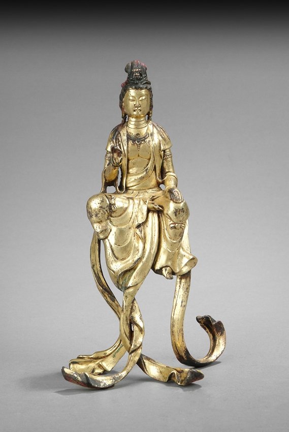 A GILT BRONZE FIGURE OF GUANYIN: Seated in Lalitasana, the Bodhisattva is dressed in loose robe draped over shoulder and left open at the chest exposing an ornamented necklace, the long scarves extends down and acting as support. 18t