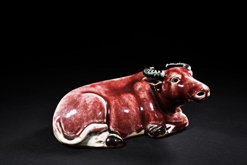 A RED-GLAZE BUFFALO: In recumbent position, the buffalo is modelled with legs tucked beneath the rounded body and tail flicked to one side, the head turned to one side, the body covered in red glaze and horns in green gla