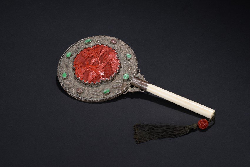AN ANTIQUE CINNABAR LACQUER BONE HAND MIRROR: The antique circular mirror back is mounted with a shaped cinnabar lacquer panel depicting a lady standing besides a lake, the back decorated with eight buddhist treasures in repousse and gems alterna