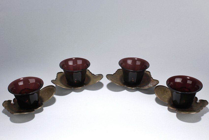 A SET OF GLASS CUPS WITH SILVER STANDS: Each is identical in design and all possess a maroon-violet hue. It rests on a short foot that gradually curves out to form the body and mouth. The cups are perfectly smooth all around an sit on top o