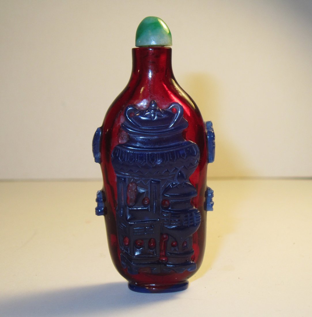 A Chinese Cobalt Blue Overlay Red Glass Snuff Bottle: Circa 1890 - 1949, Of rounded rectangular body in deep red ground overlaid with blue still life scene, jade colored glass stopper, recessed oval foot. Height 2.75 in. (7 cm)