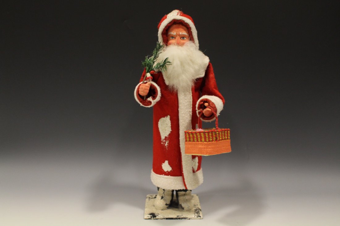 Tall Santa Claus Candy Container Holding Basket Ex Cond: Santa Claus / Father Christmas papier / paper / paper Mache candy container, wearing a long red robe holding basket and feather tree. His costume, boots and wooden base with patches of painted on snow