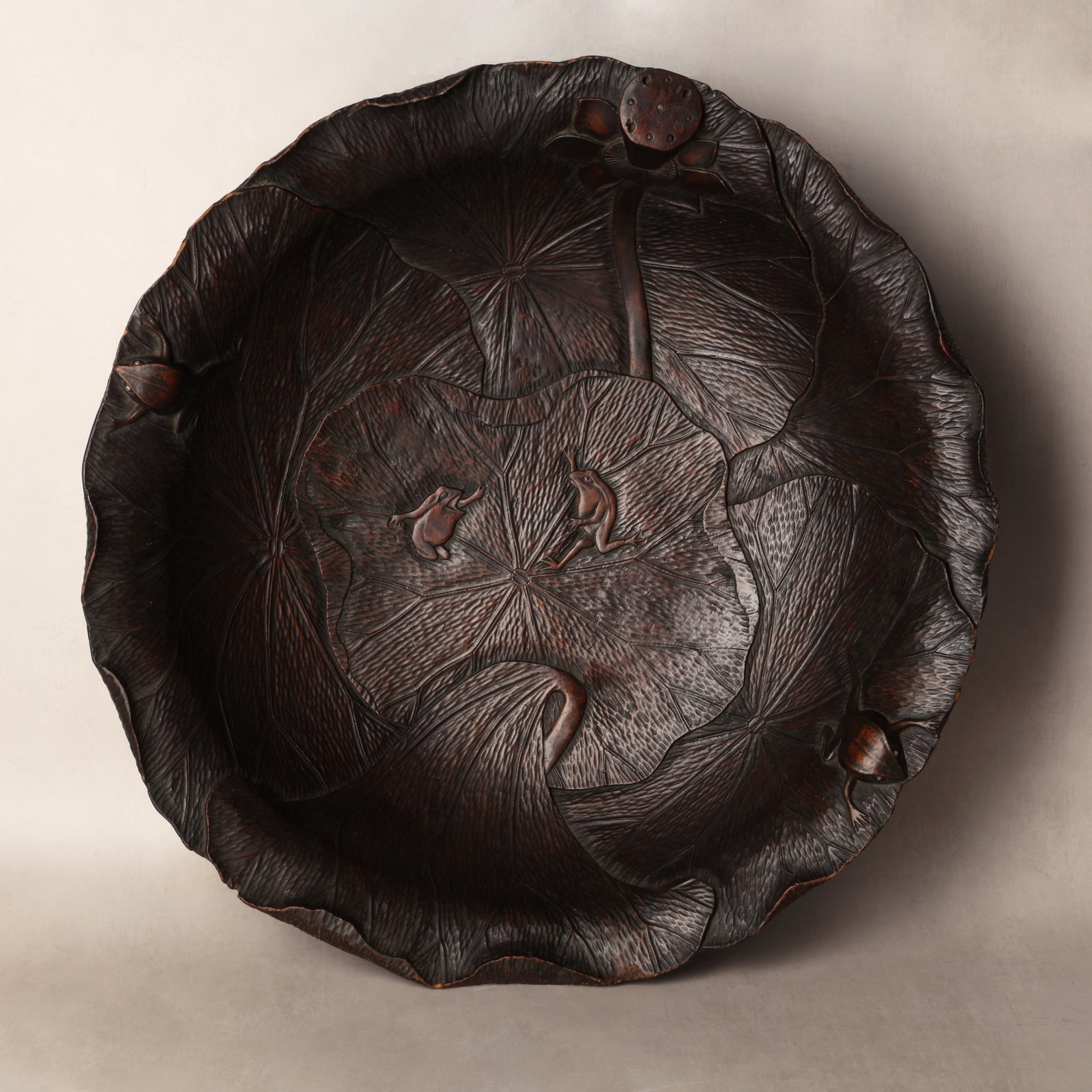 A large Japanese wooden 'lotus' bowl, Meiji period, 19th century: The robustly carved bowl of large proportions in the form of open curled lotus leaves, further adorned with frogs and lotus buds, with fine detailing for the stems and veins of the leaves Size:(60.5cm