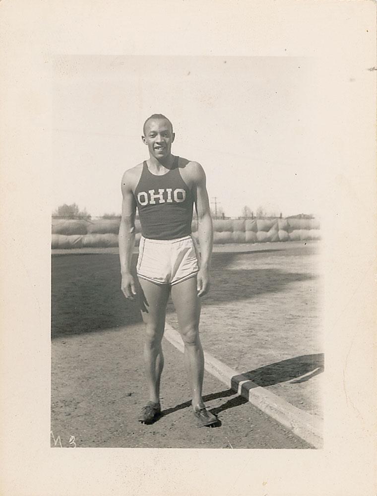 Jesse Owens: Very early vintage glossy 2.75 x 4.5 full-length photo of a young Owens in his track uniform and shoes posing on a field, signed in fountain pen. Accompanied by a second unsigned vintage photo, 3 x 4,